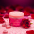 Crimson Kiss Royal Jelly body scrub in a pink jar surrounded by rose petals and sparkling decorations.